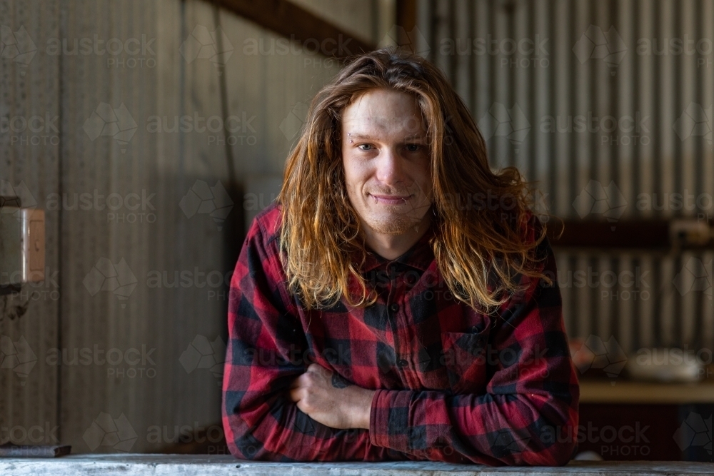 young man in flannel shirt leaning on bench in shed - Australian Stock Image