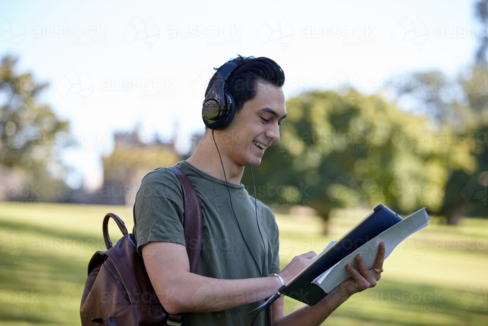 Young man laughing outside holding his device wearing headphones at park - Australian Stock Image