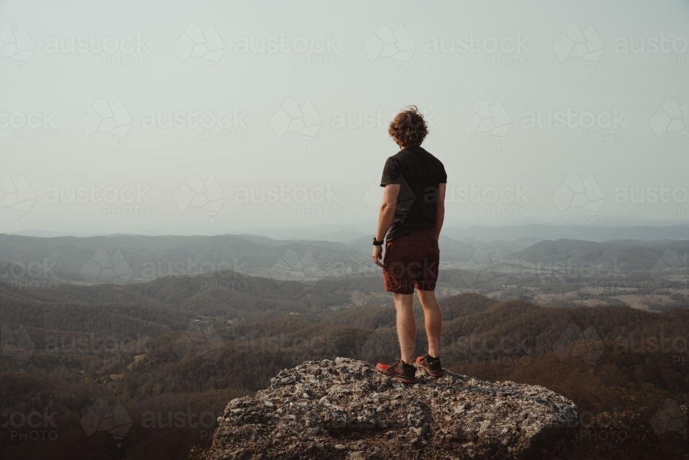 Young man on a mountain top looking at the views of the valley below. - Australian Stock Image