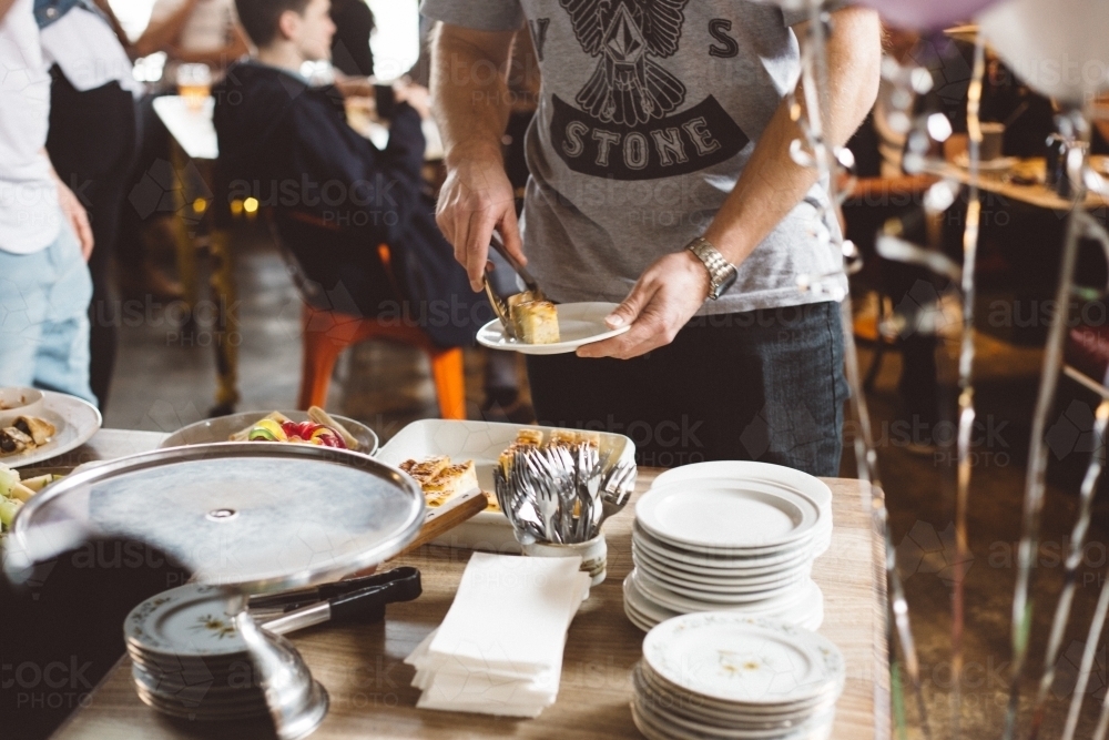 Young man putting food from a serving table on a plate, people in background at a party - Australian Stock Image