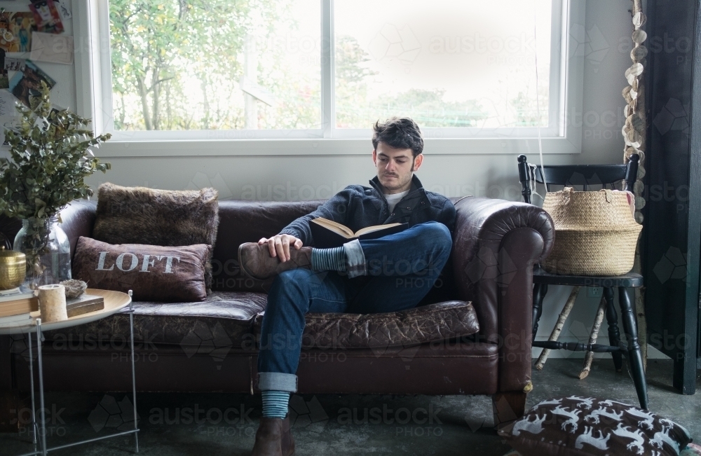 Young man reading a book on a worn leather sofa - Australian Stock Image