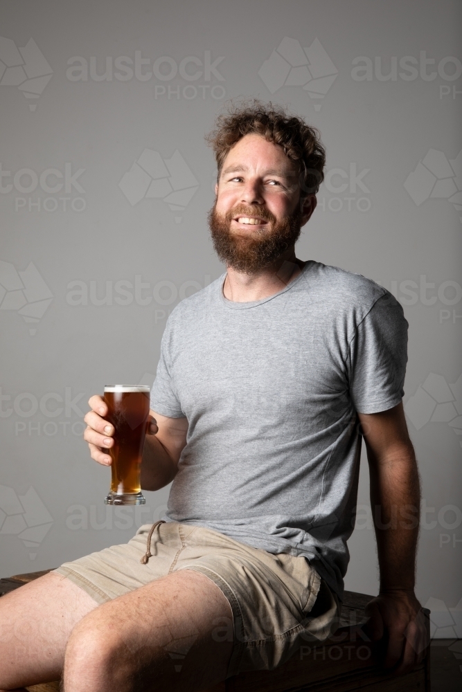 Young man sitting, holding a glass of beer, relaxed and happy - Australian Stock Image