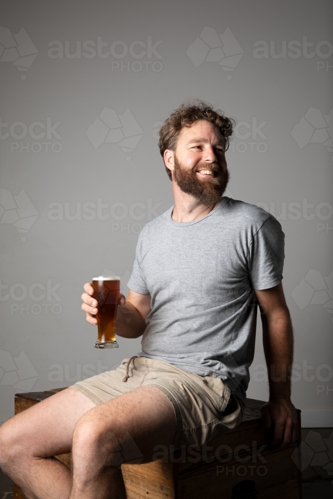 Young man sitting, holding a glass of beer, relaxed and happy - Australian Stock Image