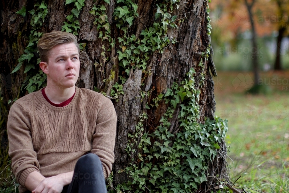 Young man sitting outdoors at the base of a tree contemplating life - Australian Stock Image