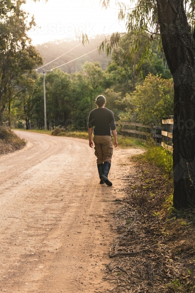 Young man walking down winding dirt road on bush walk at sunset outside rural property - Australian Stock Image
