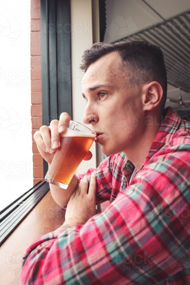 Young man wearing a flannel sipping a glass of craft beer as he stares out the bar window - Australian Stock Image