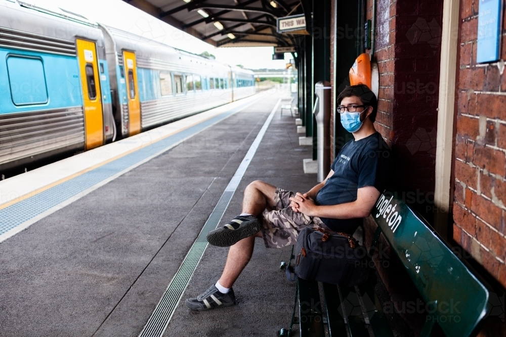 Young man wearing covid-19 face mask waiting at train station to travel to uni on public transport - Australian Stock Image