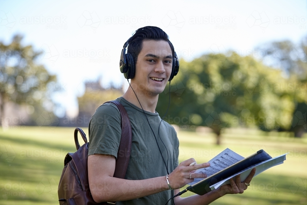 Young man with dark hair outside holding device wearing headphones at park - Australian Stock Image
