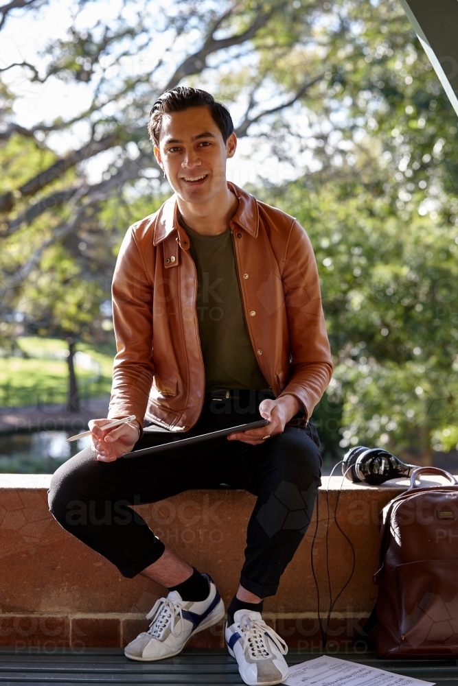 Young man with dark hair outside holding device with headphones at park - Australian Stock Image
