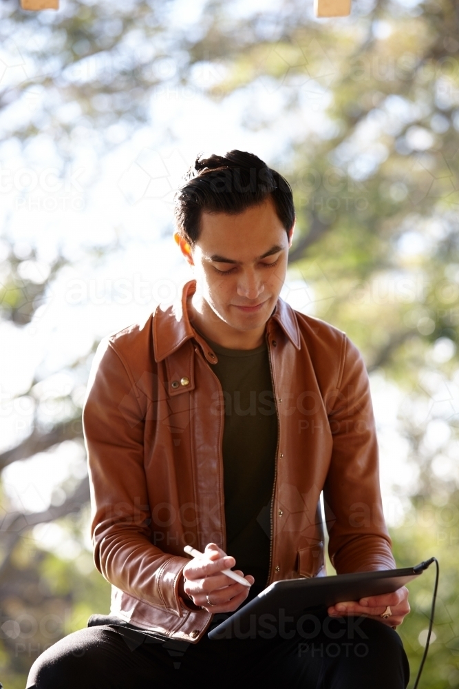Young man with dark hair outside writing on device at park - Australian Stock Image