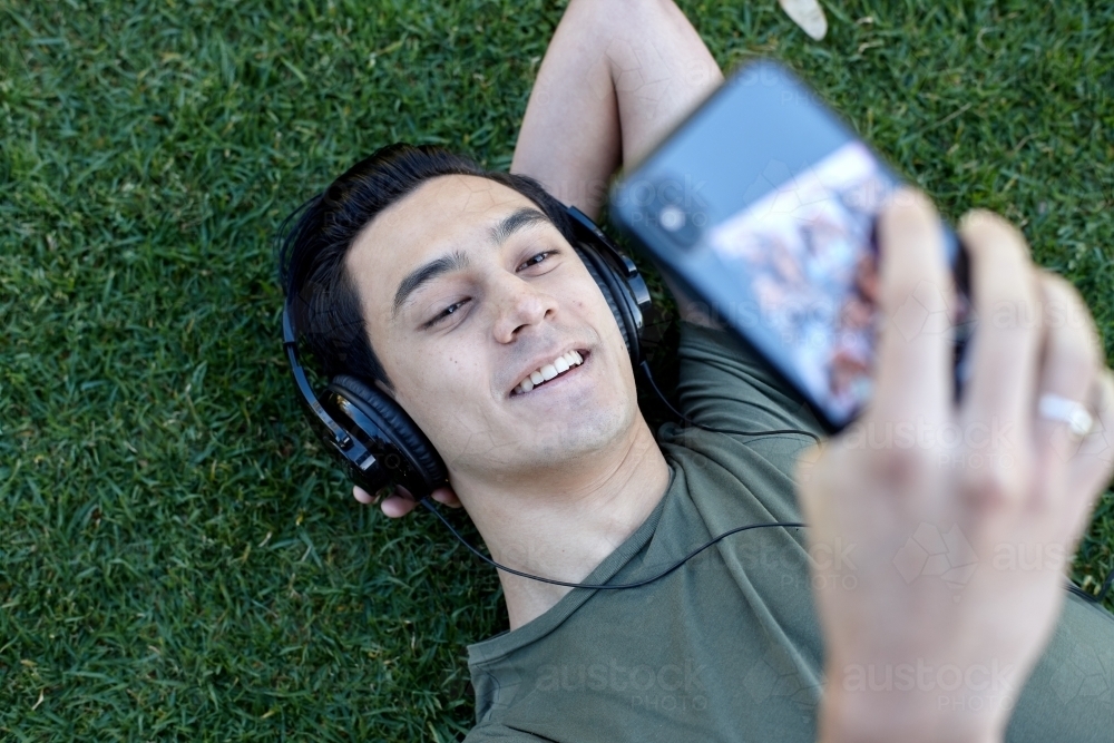 Young man with dark hair using technology on grass at park - Australian Stock Image