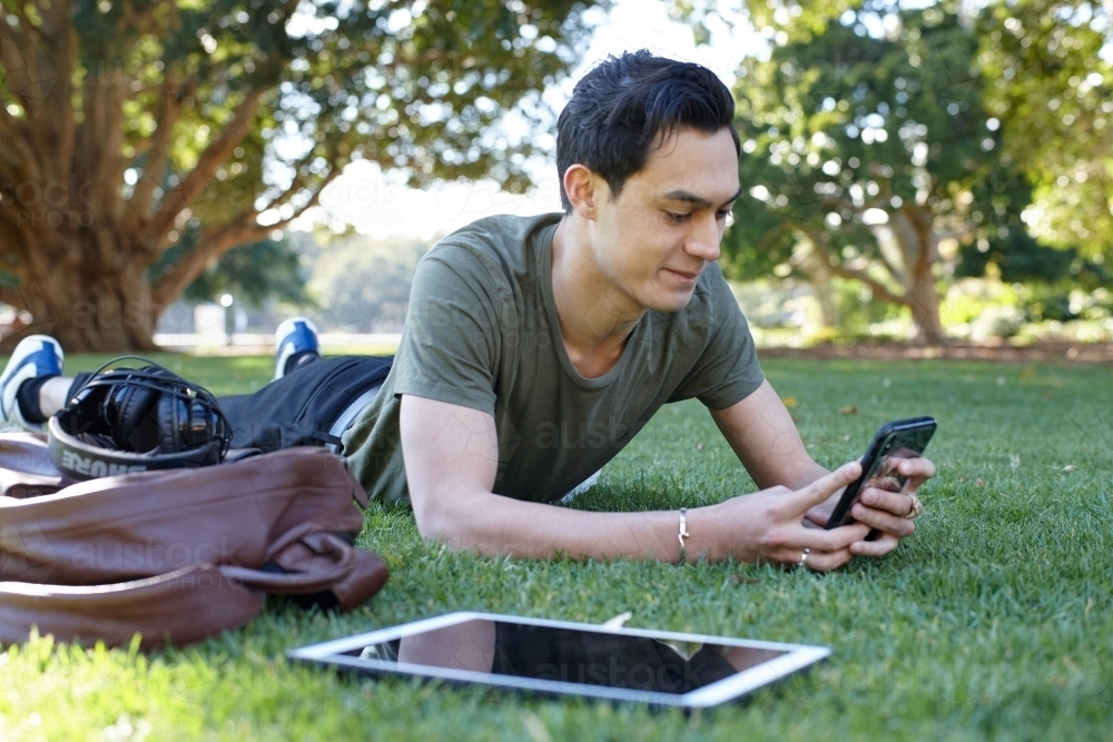 Young man with dark hair using technology on the grass at park - Australian Stock Image