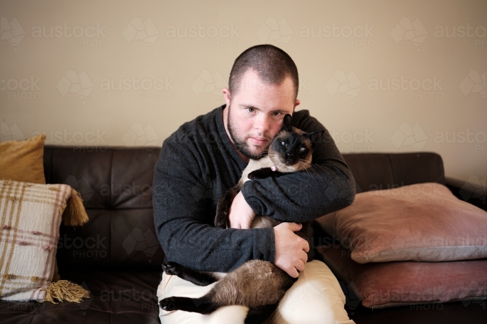 Young Man with Down Syndrome Cuddling Cat on Couch - Australian Stock Image