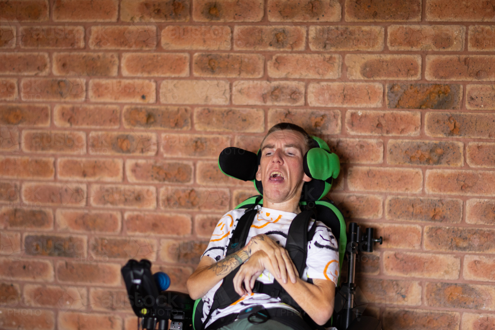 young man with tattoos on arm sitting in wheelchair in front of brick wall - Australian Stock Image