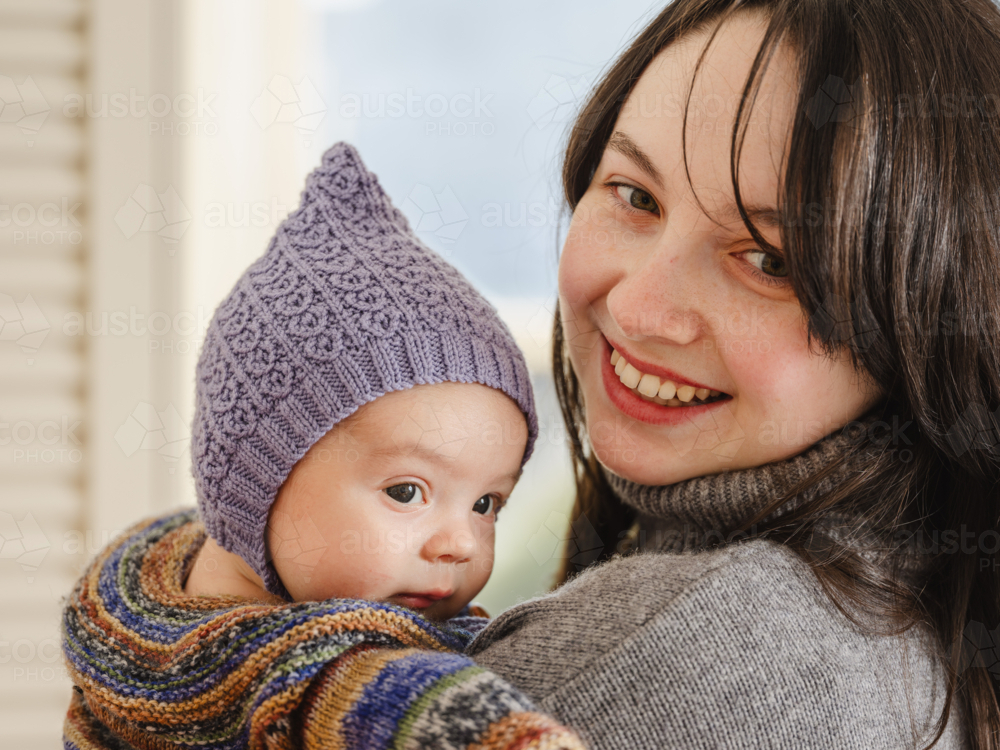 Young mother holding baby wearing beanie inside home - Australian Stock Image