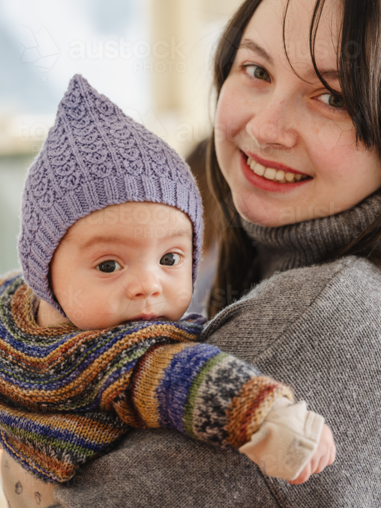 Young mother holding baby wearing beanie inside home - Australian Stock Image