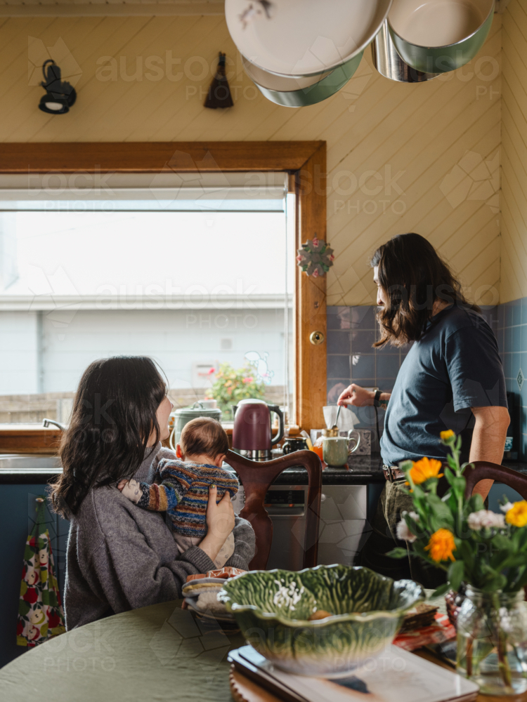 Young mother holding baby while seated and father standing drinking coffee - Australian Stock Image