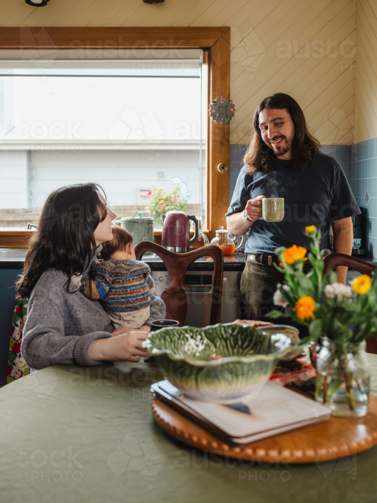 Young mother holding baby while seated and father standing drinking coffee in kitchen at home - Australian Stock Image