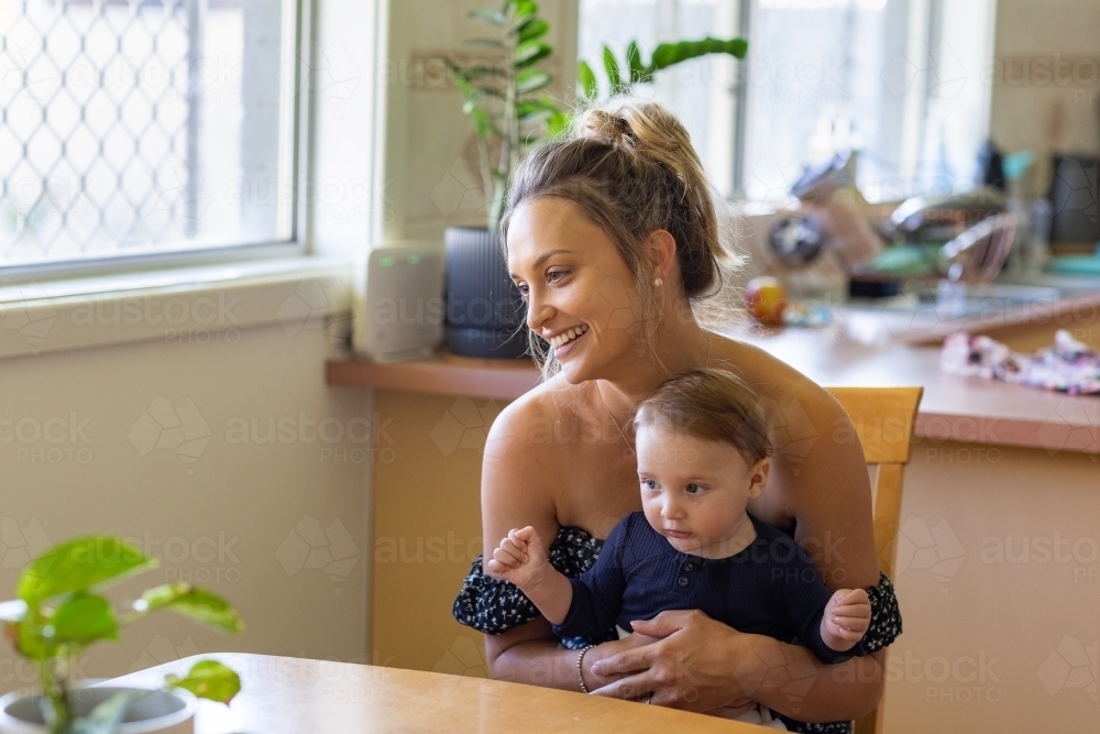 young mother sitting at kitchen table with baby on lap - Australian Stock Image