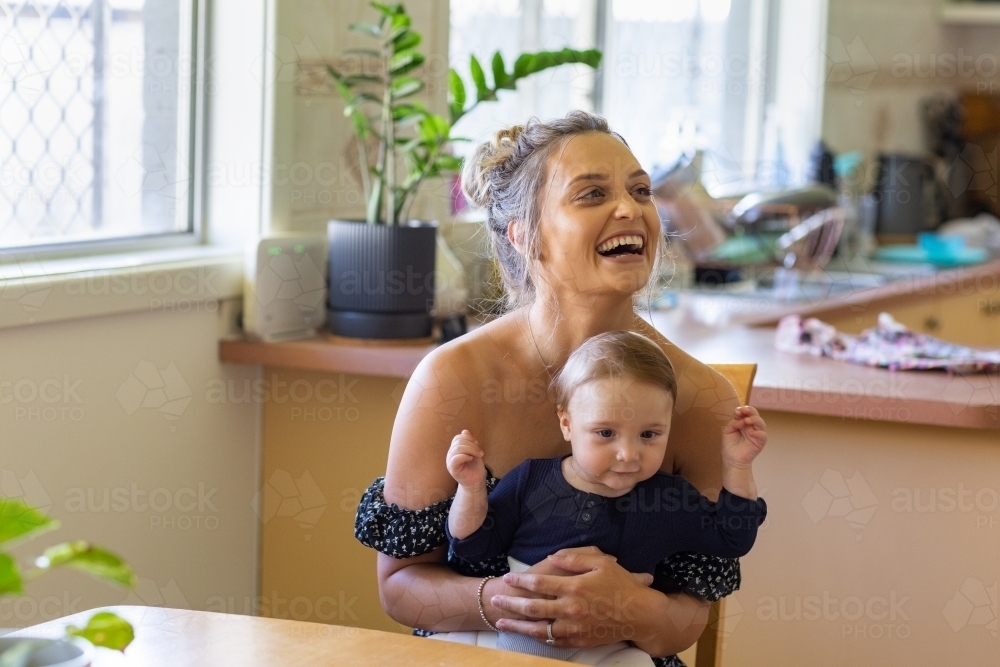 Young mother sitting at kitchen table with baby on lap - Australian Stock Image