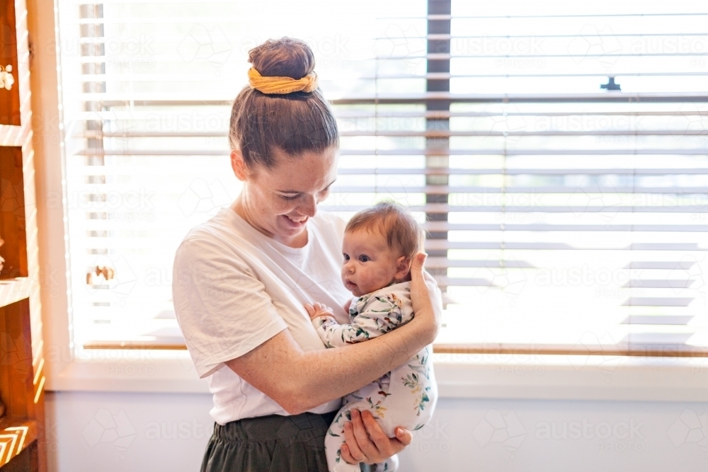 Young mother smiling and holding newborn baby inside home - Postpartum mental health - Australian Stock Image