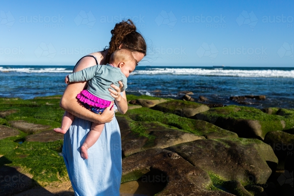 Image of Young mum carrying baby on rocks at the beach in sunlight ...