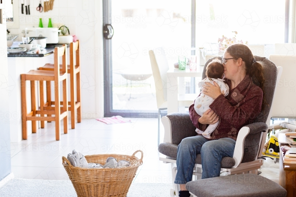 Young mum in fourth trimester singing newborn baby to sleep on rocking chair in messy house - Australian Stock Image