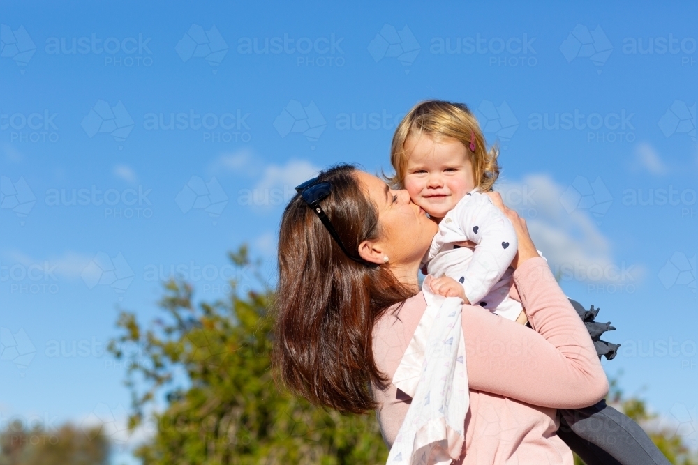 young mum lifting up and kissing toddler in front of blue sky - Australian Stock Image