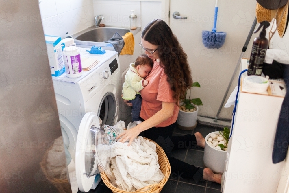 Young mum taking wet clean clothes out of washing machine while holding newborn baby - Australian Stock Image