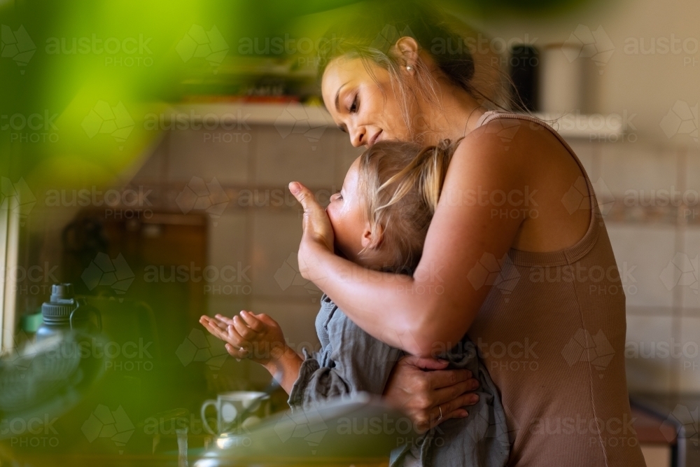 young mum wiping child's face at kitchen sink glimpsed through blurred greenery - Australian Stock Image
