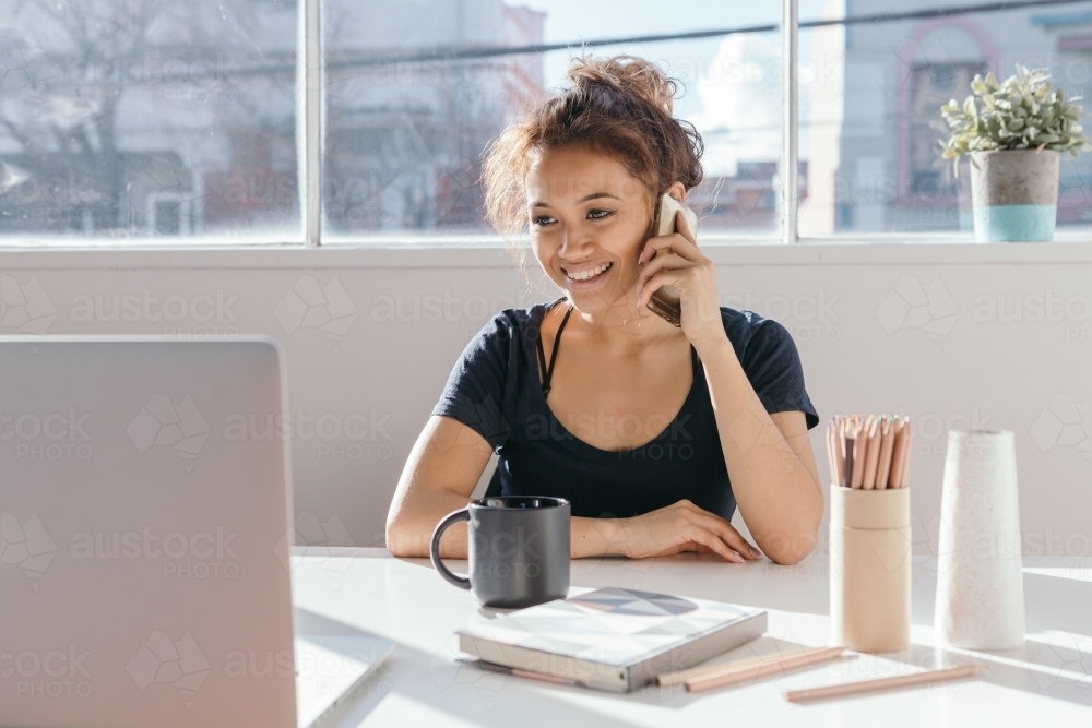 Young office employee on a mobile phone at her desk - Australian Stock Image