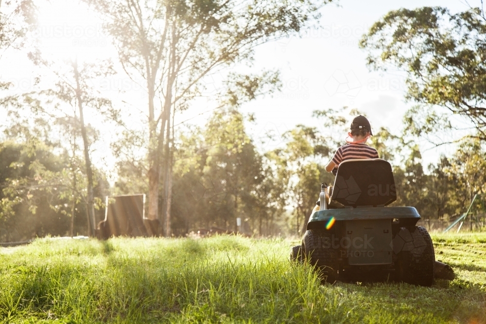 Young person mowing the paddock on a ride on lawn mower in afternoon sunlight - Australian Stock Image