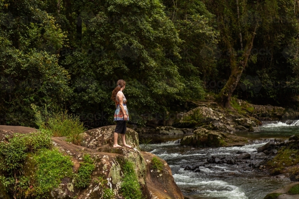 Young person standing on rock beside river in forest - Australian Stock Image
