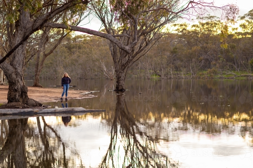 young person wearing jeans standing near a lake with trees reflecting in the water - Australian Stock Image