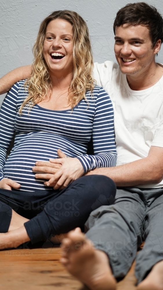 Young pregnant couple sitting against white wall - Australian Stock Image