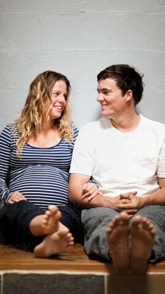 Young pregnant couple sitting against white wall, looking at each other - Australian Stock Image
