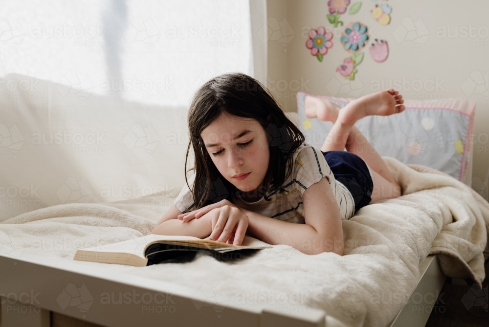 Young preteen girl in her bedroom lying on her bed reading a book by a sunny window - Australian Stock Image