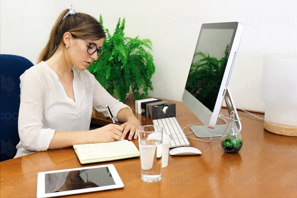 Image of Young professional woman writing in a notebook at office desk - Austockphoto