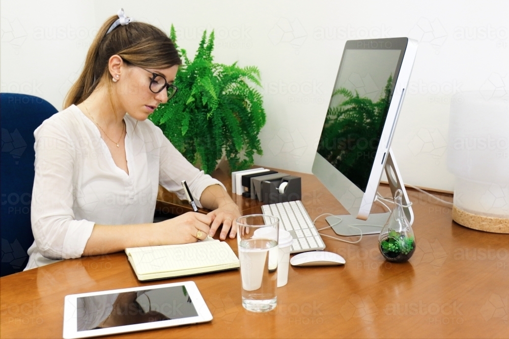 Young professional woman writing in a notebook at office desk - Australian Stock Image