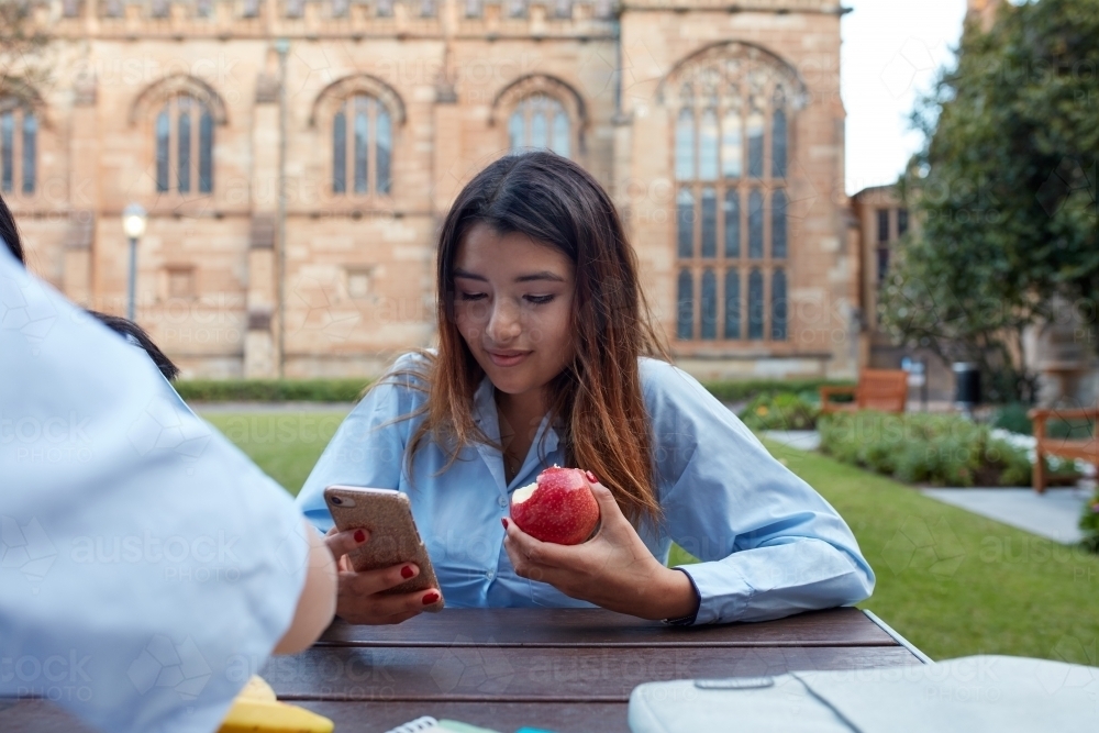 Young school girl eating apple whilst checking phone - Australian Stock Image