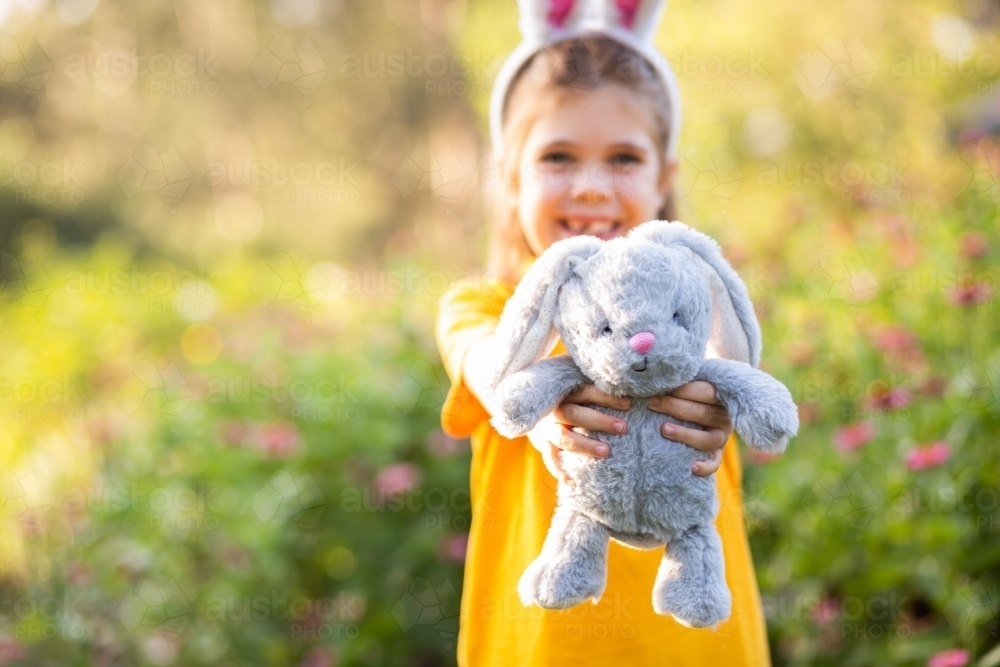 Young school girl standing outside in garden at Easter time holding out her bunny rabbit toy - Australian Stock Image