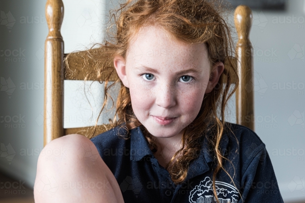 young school girl with red hair and blue eyes sitting in dining chair - Australian Stock Image