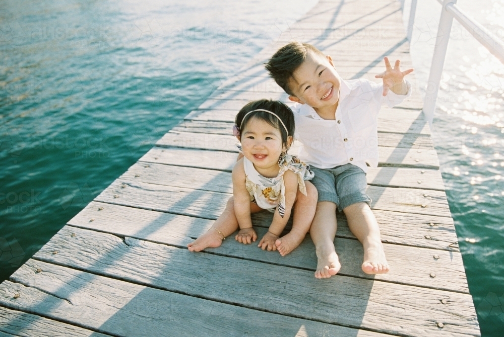 Young sister and brother sitting on wharf and smiling at the camera - Australian Stock Image