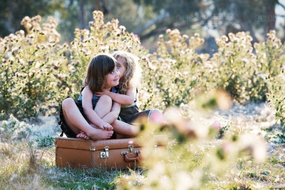 Young sisters sitting on a suitcase hugging - Australian Stock Image