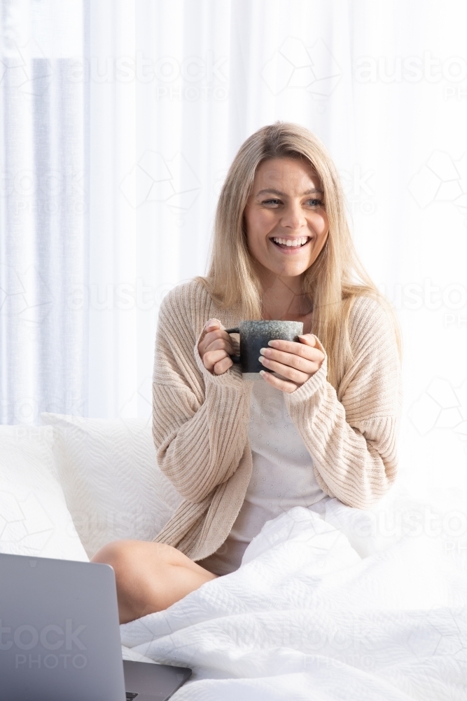 Young smiling lady drinking hot drink in white bed - Australian Stock Image