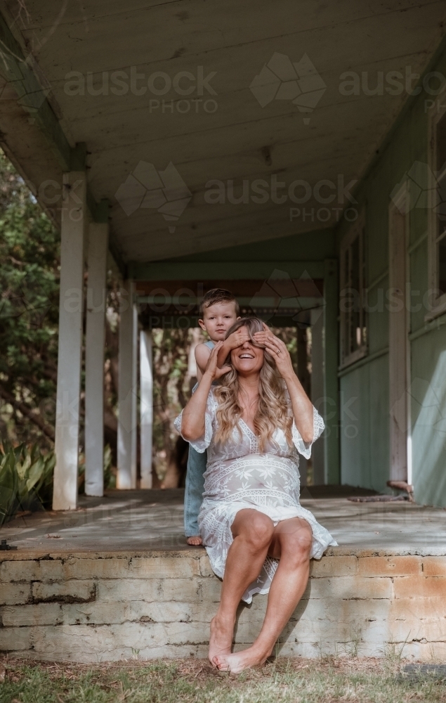 Young son covering his mother's eyes and playing with her on the front porch of home - Australian Stock Image