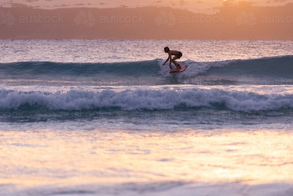 young surfer backlit surfing small waves at sunset - Australian Stock Image