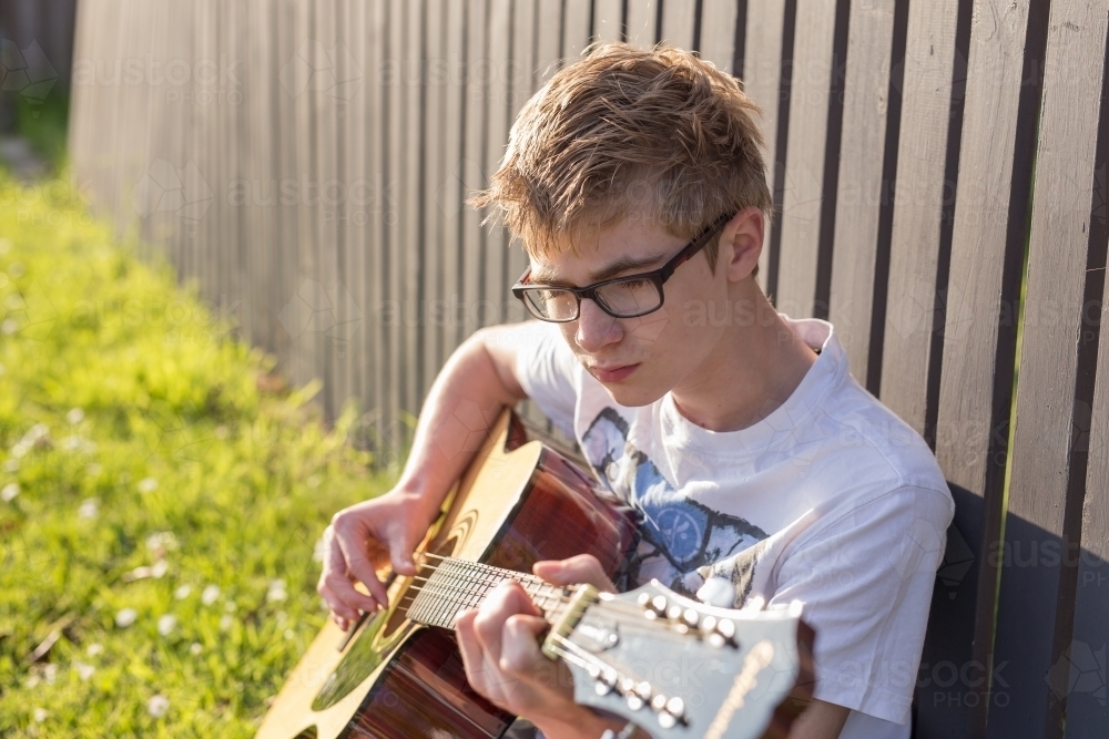 Young teen boy playing acoustic guitar by fence - Australian Stock Image