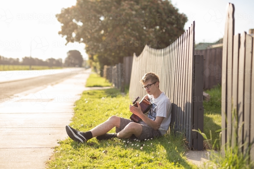 Young teen boy playing acoustic guitar by fence - Australian Stock Image