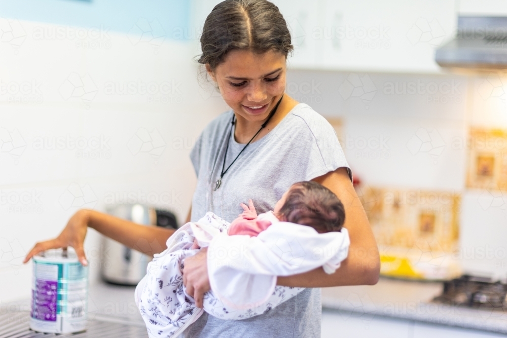 young teen girl holding her baby sister in kitchen with a can of infant formula on bench - Australian Stock Image